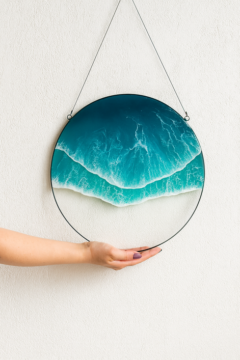 Hand holding a round ocean wave acrylic wall art with turquoise water and white foam, hanging against a textured white wall.